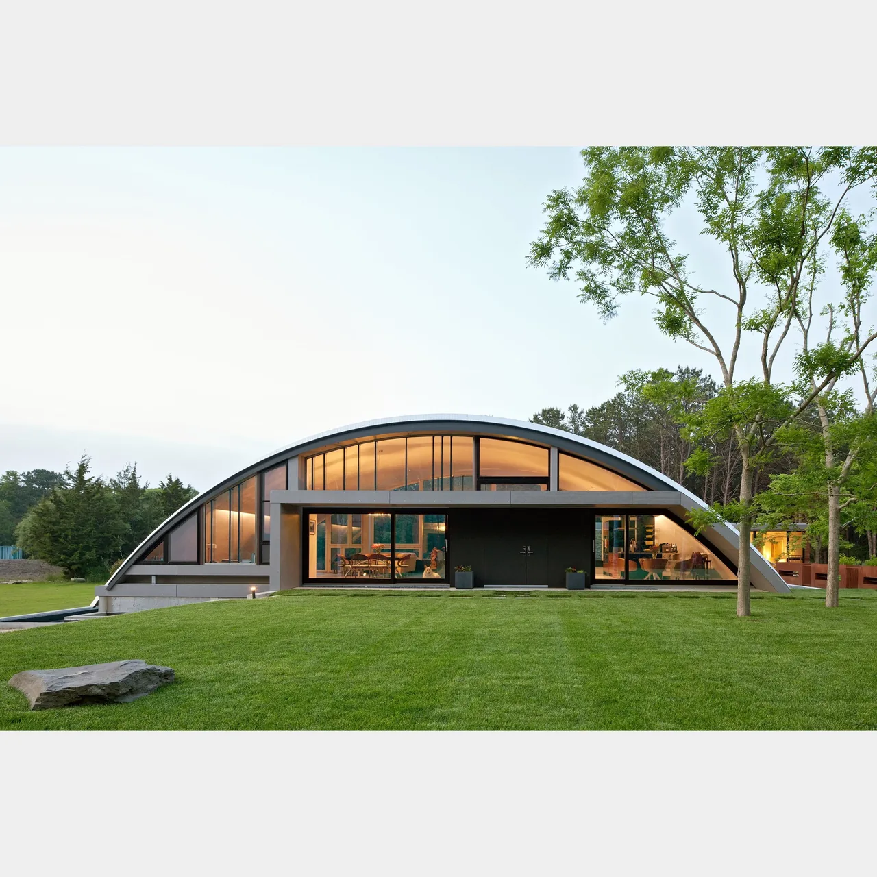 A curved modern house with large windows and a flat roof sits on a green lawn surrounded by trees.