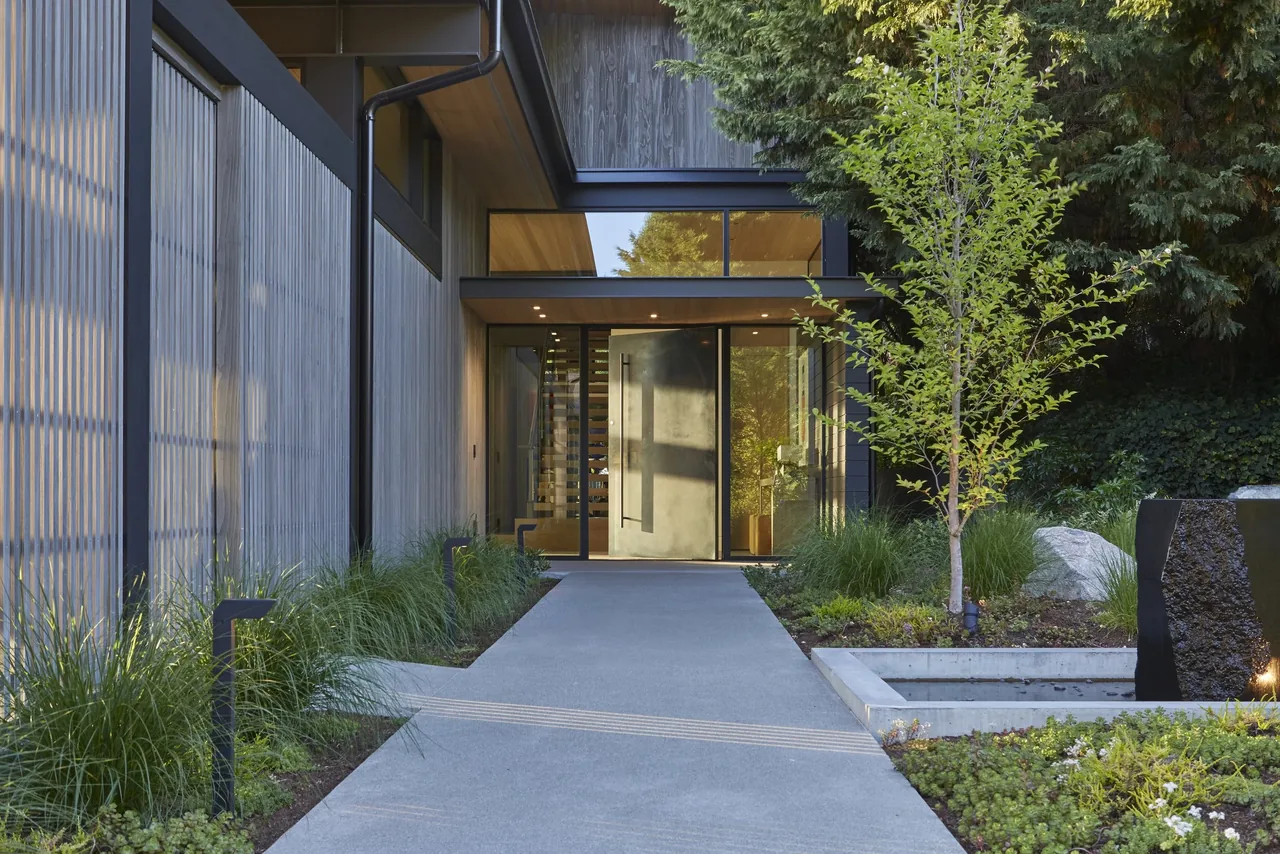 Modern house entry with large glass and metal door, surrounded by greenery and concrete pathway.