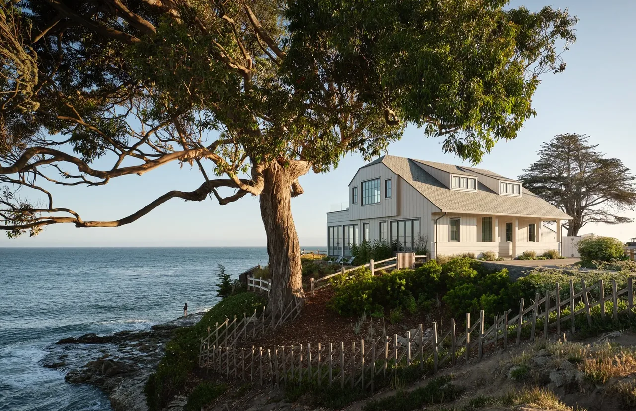 White coastal house with gray roof overlooking the ocean framed by a large tree and wooden fence.