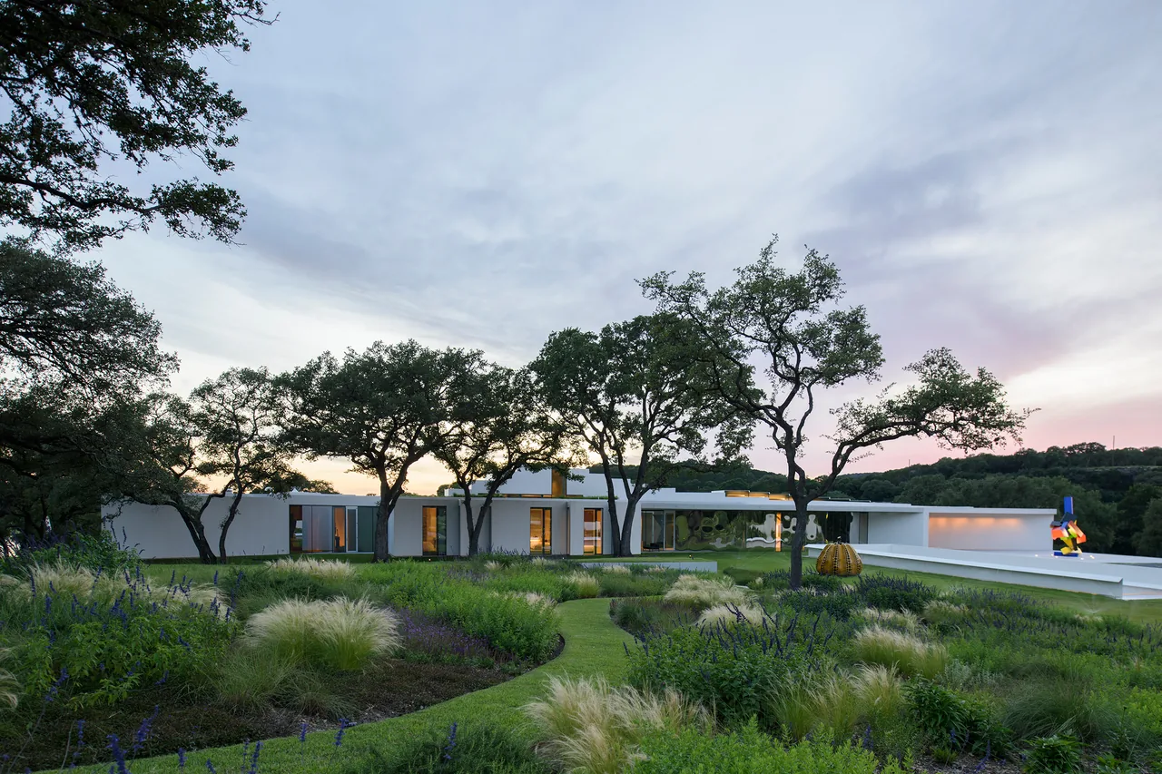 Wide view of a modern white ranch house with flat roof situated among trees and landscaped gardens during dusk.