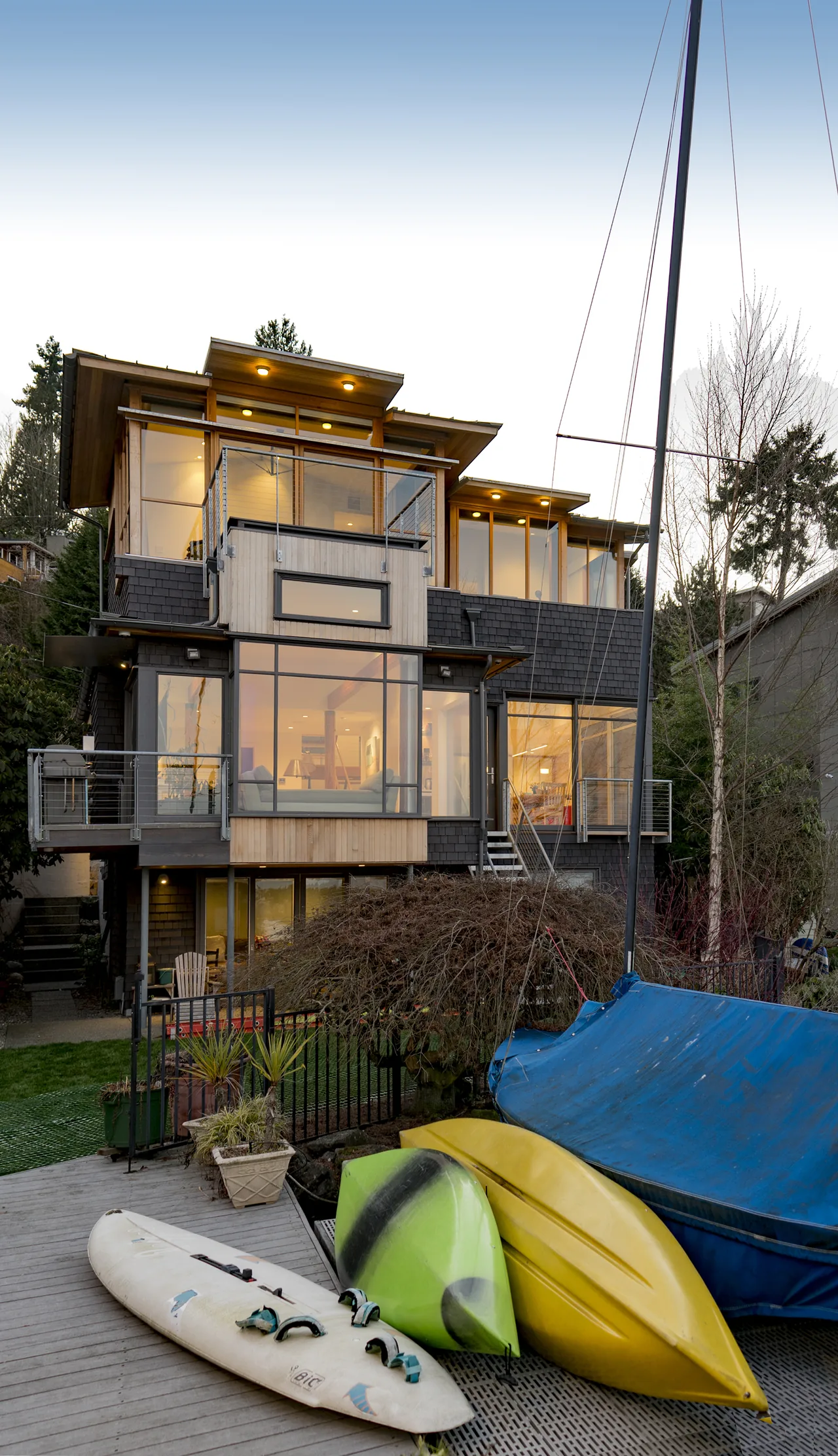 Three-story modern house with large glass windows and wooden decks, viewed from a dock with kayaks and a sailboat in front.