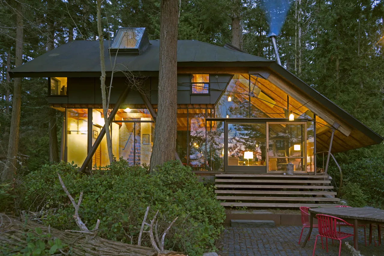 Exterior view of a glass cabin in the forest with illuminated interior and a sloped roof, surrounded by trees and outdoor seating.