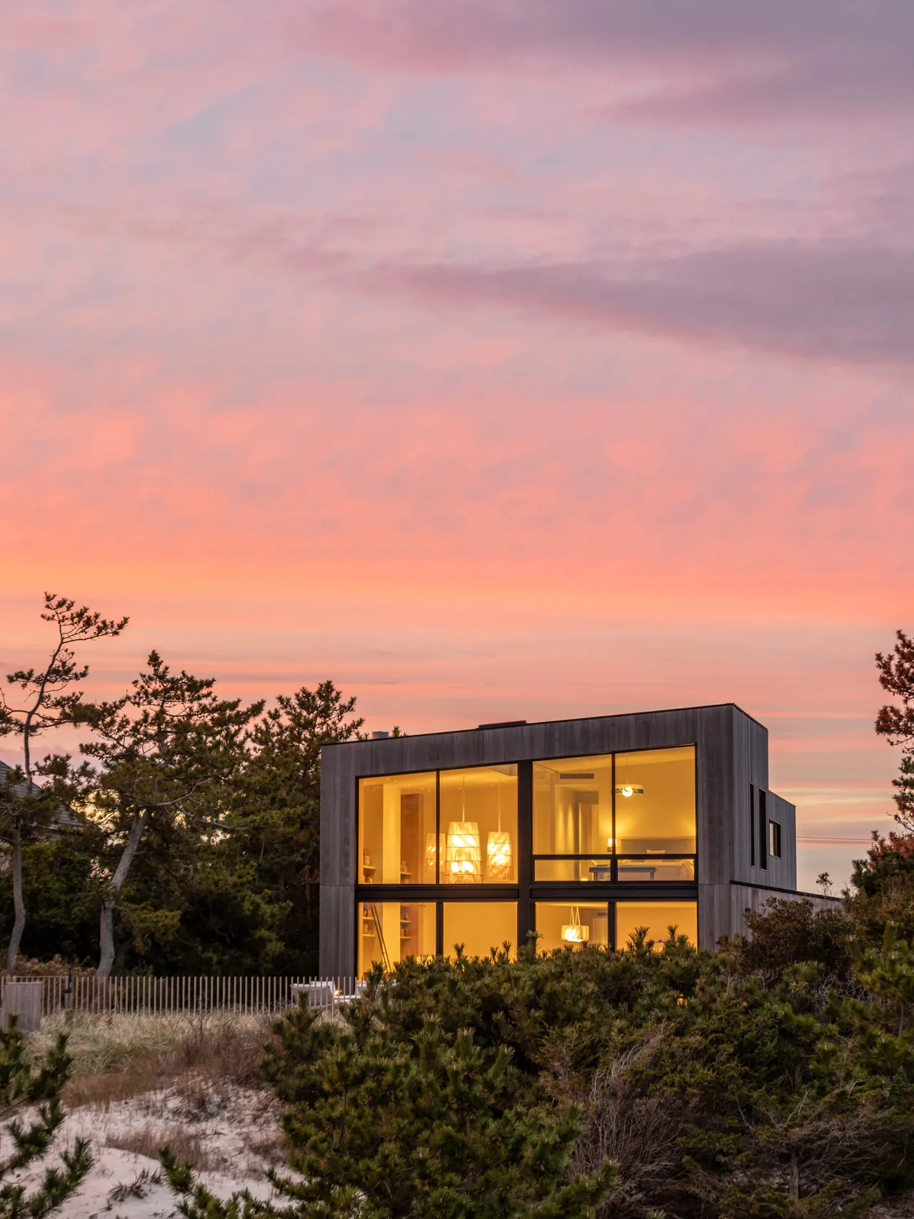 Modern two-story beach house with large floor-to-ceiling windows glowing warmly at sunset surrounded by natural vegetation.