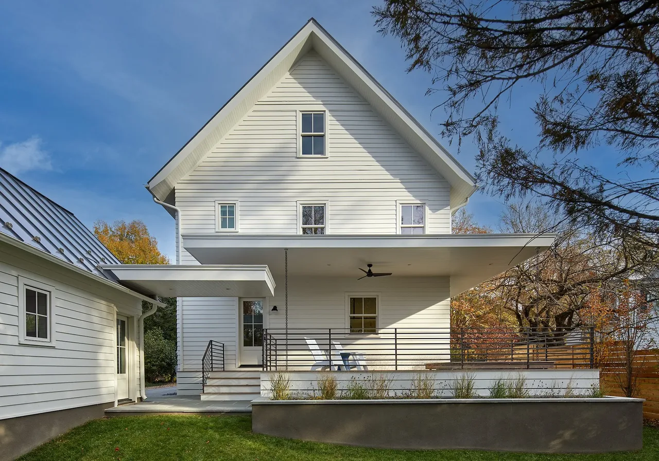 Rear view of a white house with gable roof, a covered patio with ceiling fan, and horizontal metal railing seating area.