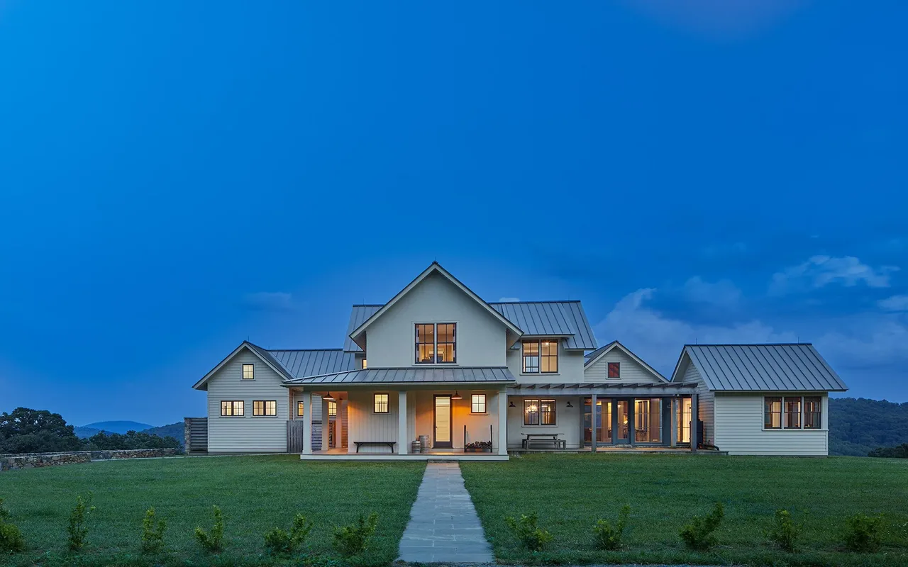 Front exterior view of a modern farmhouse residence at dusk with metal gable roofs and white siding, set on a large lawn.