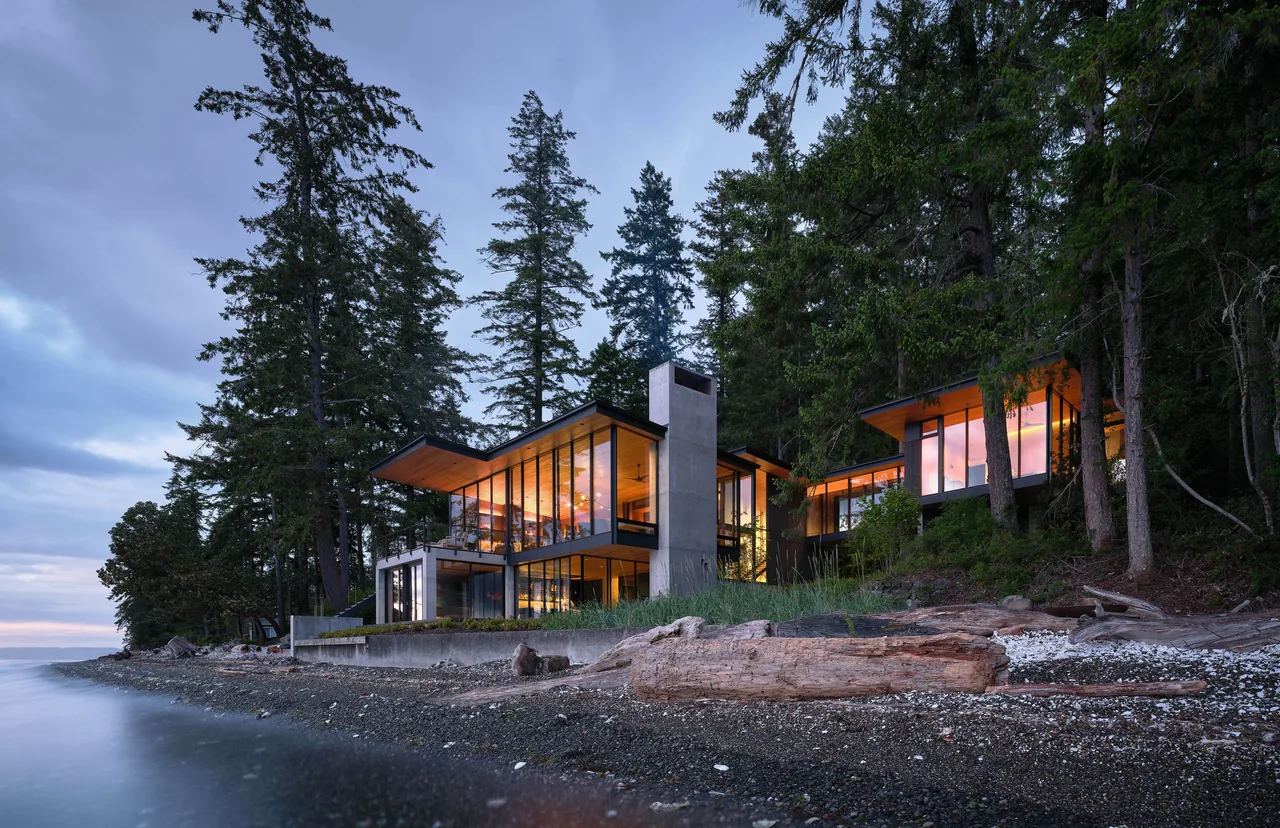Modern lakeside house at dusk with large glass windows and overhanging flat roofs, surrounded by tall evergreen trees.