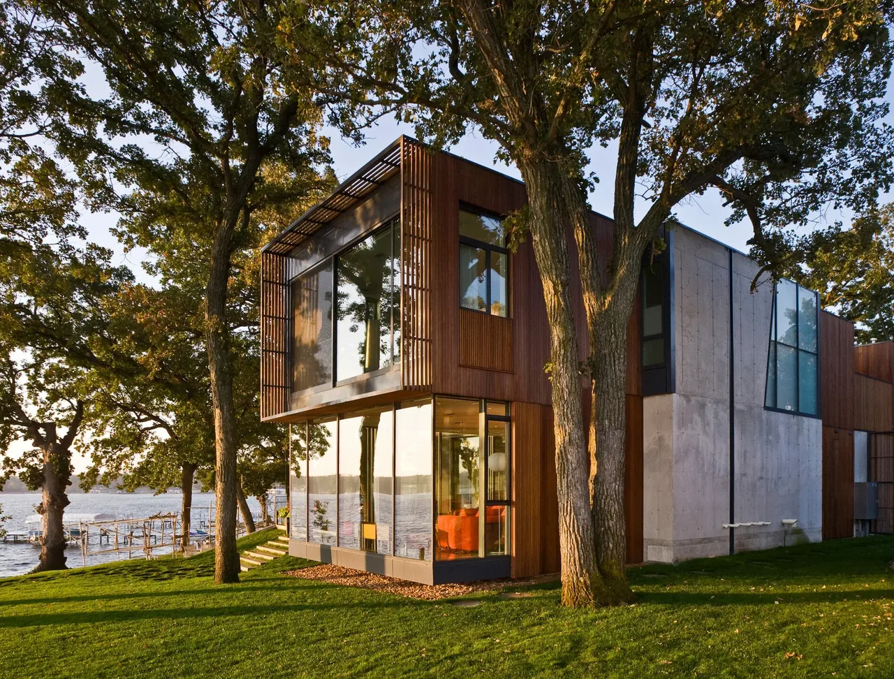 Modern two-story house by a lake with large floor-to-ceiling windows and wood and concrete exterior walls.