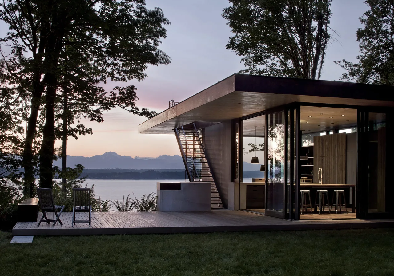 Modern house exterior at dusk overlooking water with large glass walls and outdoor seating on a wood deck.