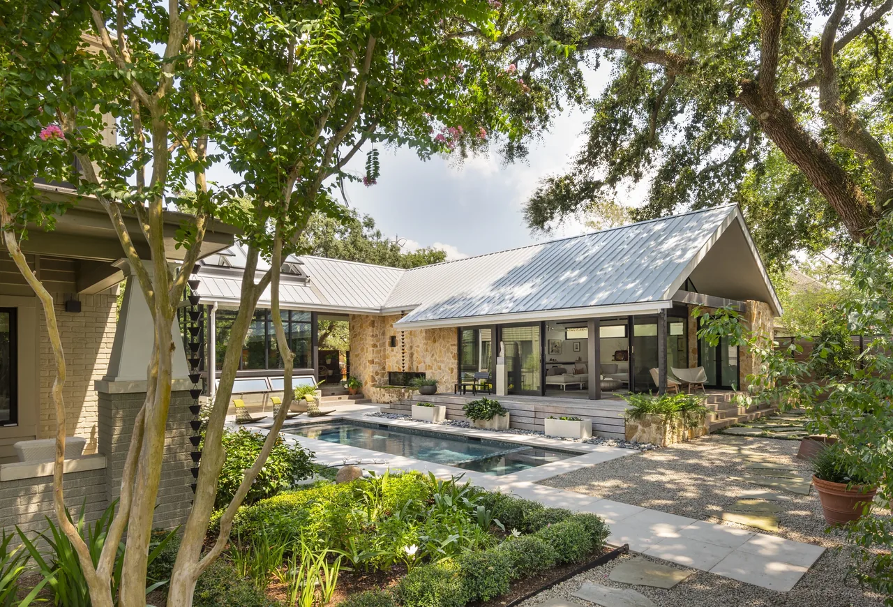 Outdoor view of a garden house with stone walls, large glass windows, and a metal gable roof surrounding a rectangular pool with lush green;