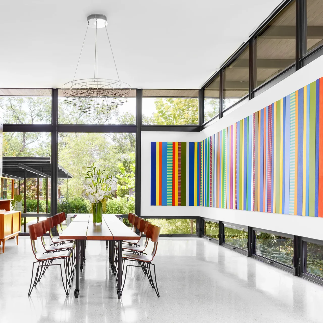 Bright dining room with large wall art, wood table, modern pendant light, and floor-to-ceiling windows overlooking greenery.