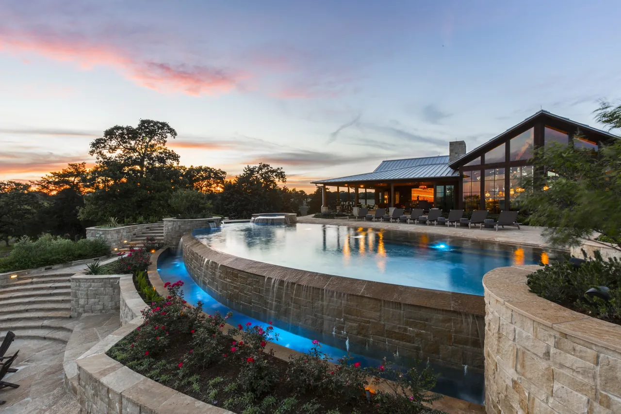 Outdoor nighttime view of a modern ranch house with a large curved infinity pool surrounded by stone walls and lounge chairs.