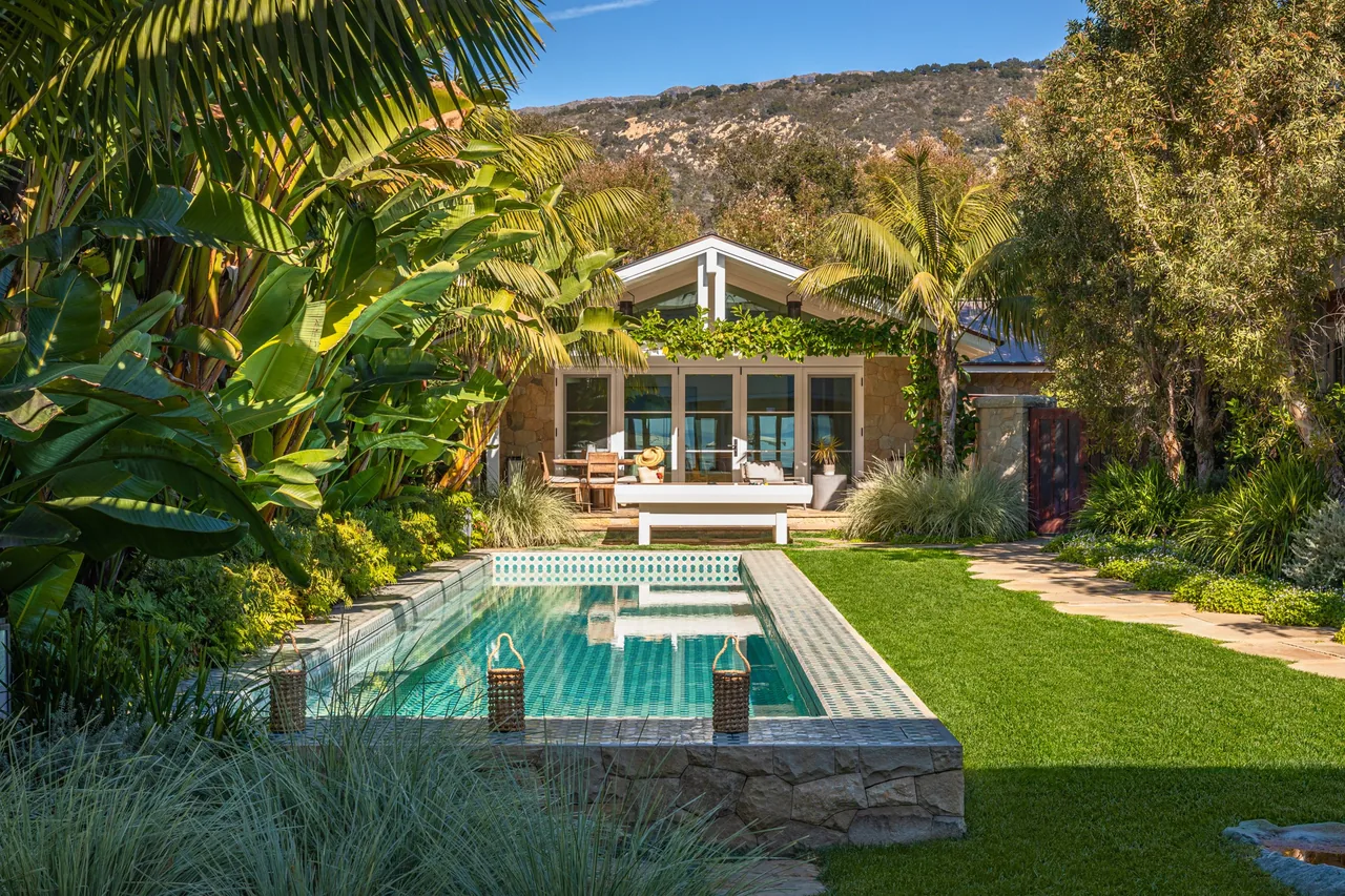 A backyard with a rectangular swimming pool surrounded by tropical plants and a house with a gable roof in the background.
