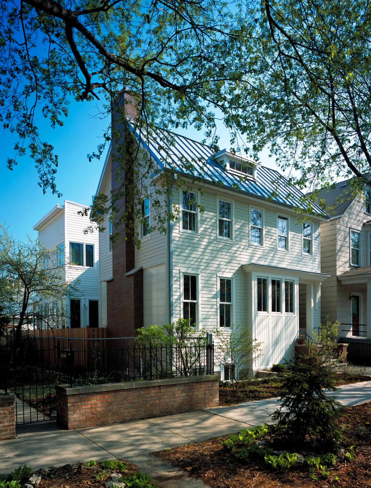Two-story house with white siding, brick chimney, metal gable roof, and surrounded by trees and garden.