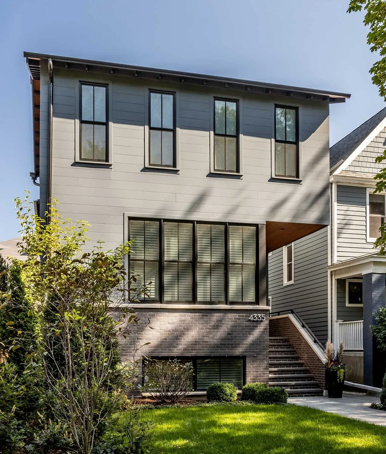 Front exterior of two-story modern house with gray siding, black-framed windows, and brick base, surrounded by greenery.