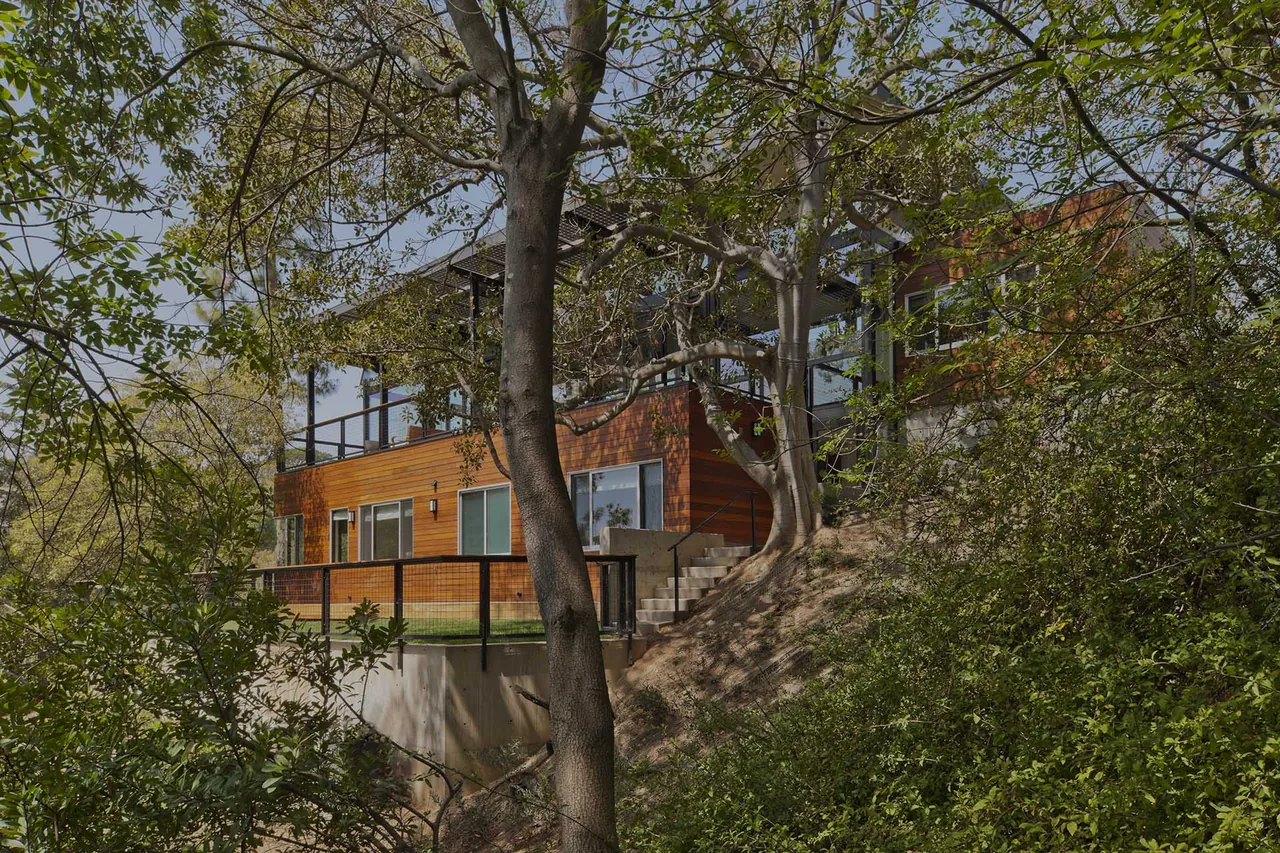 Modern two-story house with wood siding situated on a hillside surrounded by trees.