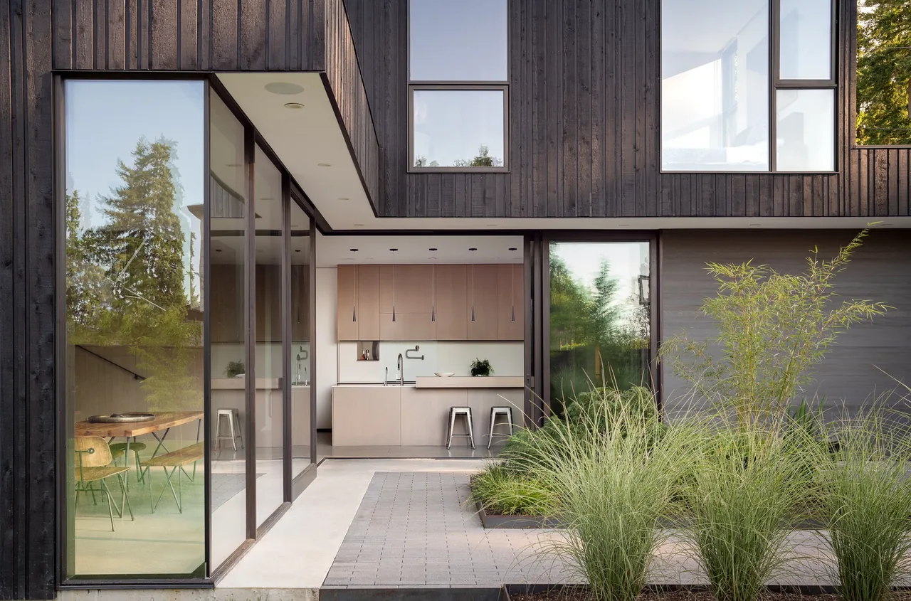 Modern courtyard with black wood siding exterior, large glass windows, and a view into a minimal kitchen area.
