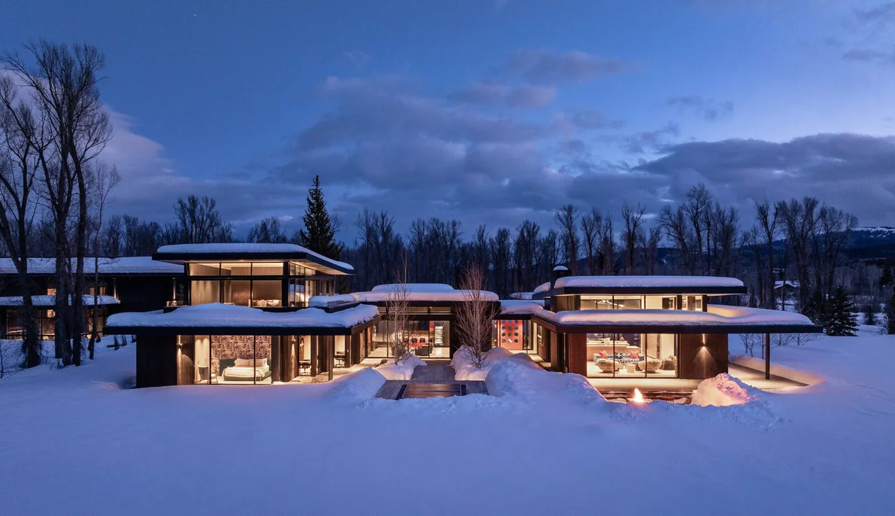 Modern multi-wing house covered in snow with extensive glazing and warm interior lighting at dusk.