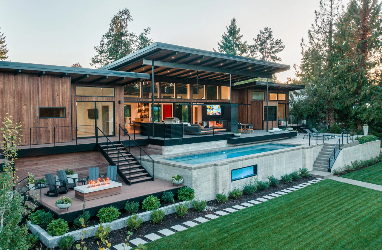 Modern wooden house with large glass windows, covered patio, outdoor pool, and fire pit with seating in a landscaped yard.