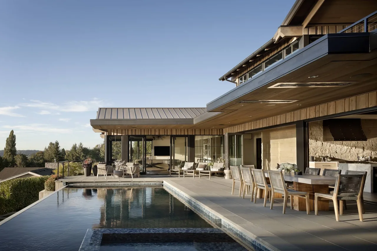 Modern outdoor living area with pool, dining table, and lounge chairs under a canopy roof extending from the house.