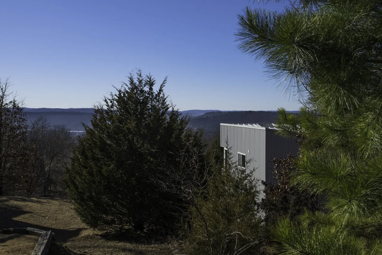 View of a metallic-sided modern building partially obscured by trees on a hillside with distant hills under a clear blue sky.