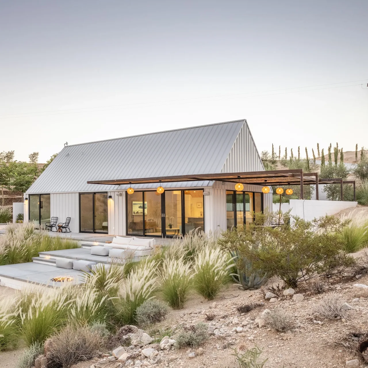 Small white guesthouse with a steep metal gable roof and glass sliding doors opening to outdoor seating and a pergola with hanging lights.