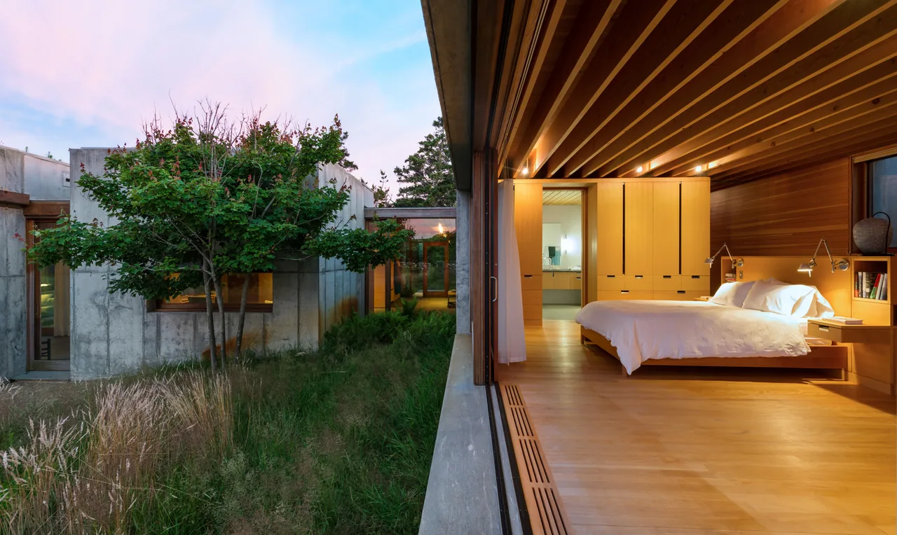 View of a bedroom interior with warm wood paneling and a bed, opening onto a courtyard with concrete walls and a tree.