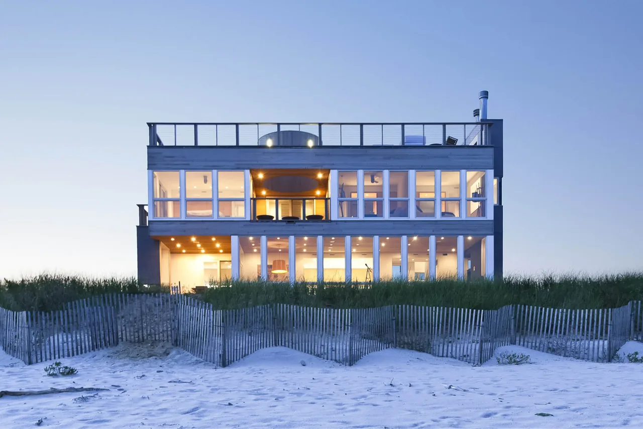 View of a modern two-story beach house at dusk with large glass windows and outdoor lighting.