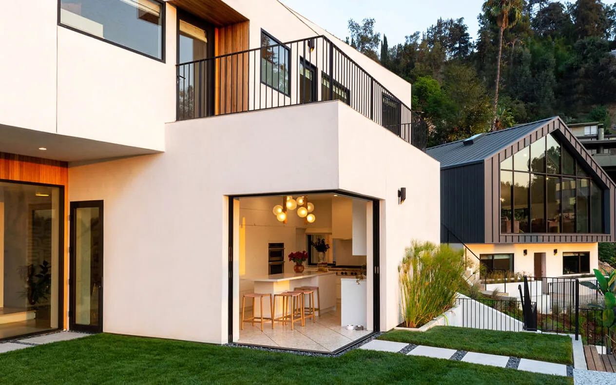 Exterior view of a modern hillside home with large corner windows and a kitchen visible inside.