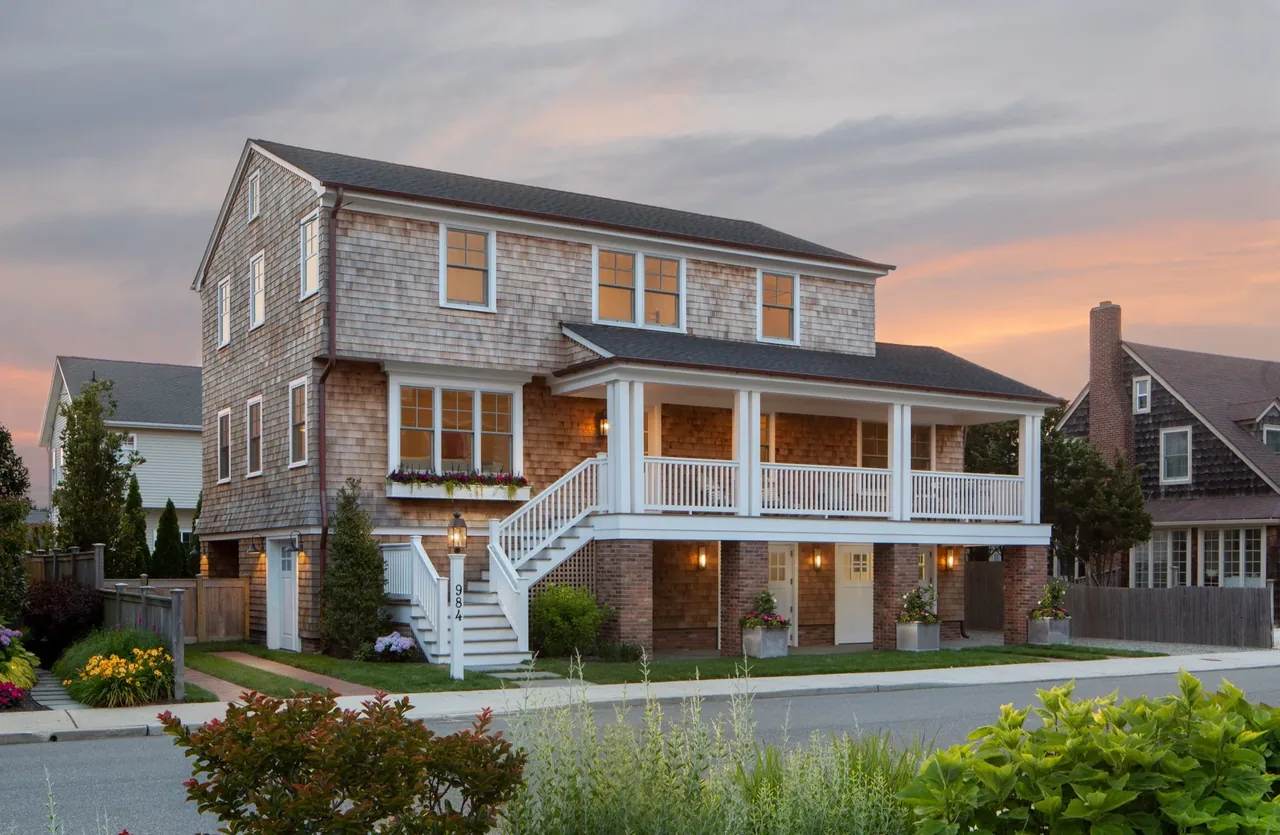 Two-story house with shingle siding and a large covered porch raised on brick columns at sunset.
