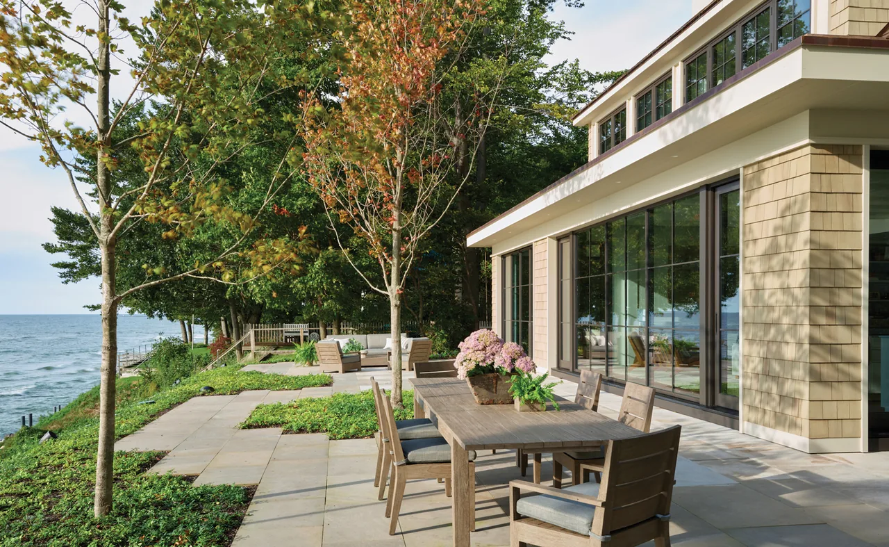 Patio dining area with wood table and cushioned chairs beside large windows overlooking a lakeside landscape with trees.