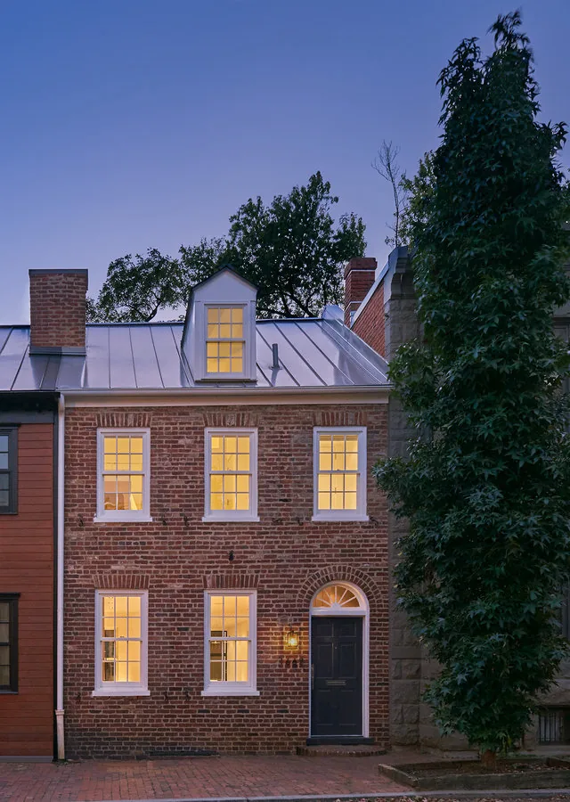 Brick townhouse with lit windows at dusk and a metal gable roof with a dormer window.