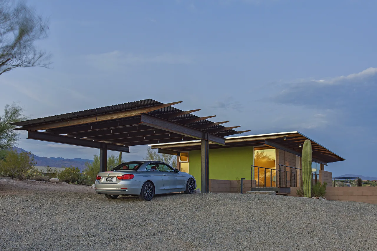 Photo of a modern house exterior in a desert landscape featuring a carport with exposed steel beams and a corrugated metal roof.