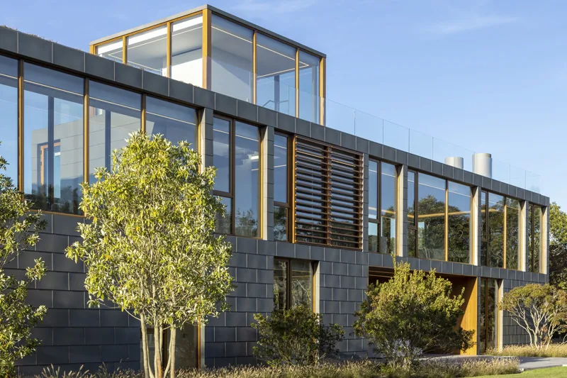Exterior view of a modern two-story house with large glass windows and metal cladding surrounded by small trees and shrubs.
