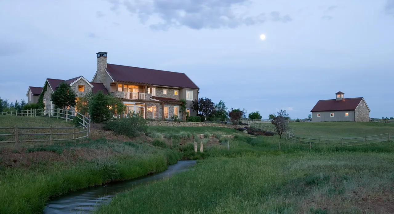 Stone farmhouse with red metal gable roofs and a chimney, next to a small barn and fenced grassy landscape under a cloudy sky at dusk.