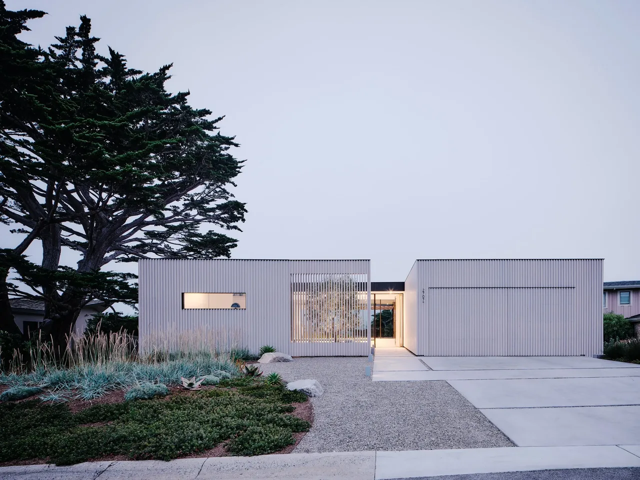 Modern flat-roofed house with linear windows and vertical siding, lit in early evening with landscaped gravel driveway.