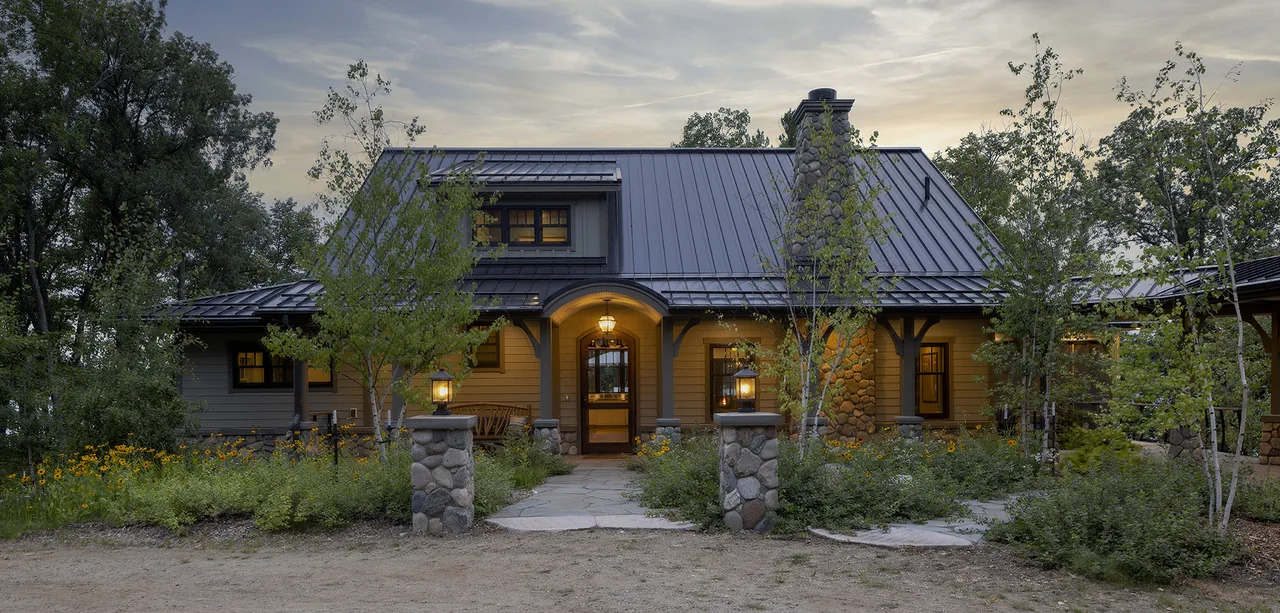 Exterior front view of a lodge-style house with stone pillars, a metal roof, and a large stone chimney framed by trees.