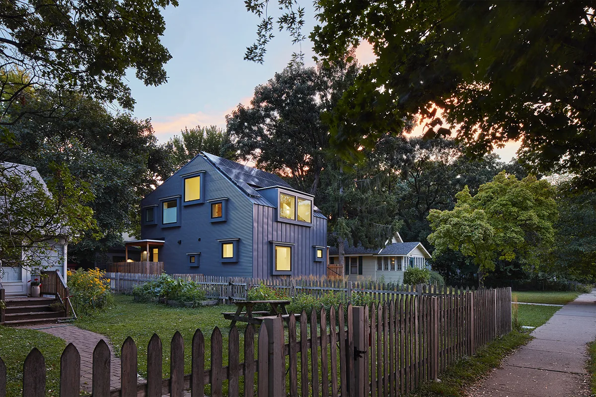Exterior view of a modern dark blue two-story house with multiple windows illuminated at dusk, surrounded by trees and a wooden picket fence