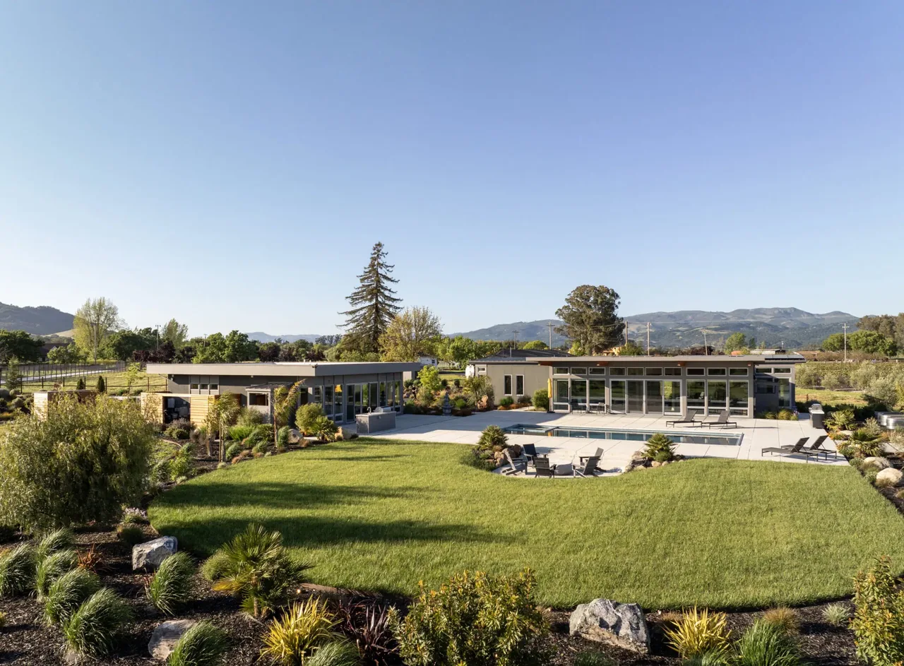 Modern single-story home with flat roofs and large glass windows overlooking a landscaped lawn with a pool and lounge chairs.