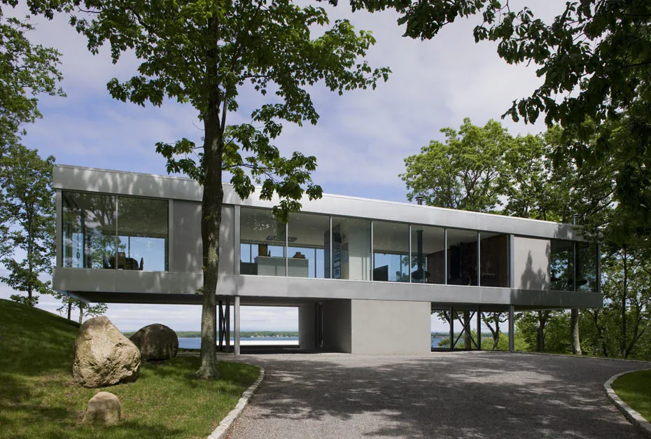 Modern elevated house with floor-to-ceiling glass windows surrounded by trees and rocks.