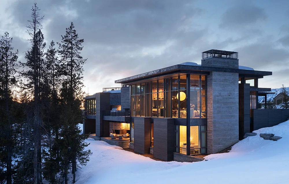 Modern multi-level house with large floor-to-ceiling windows and stone chimney at dusk in snowy landscape.