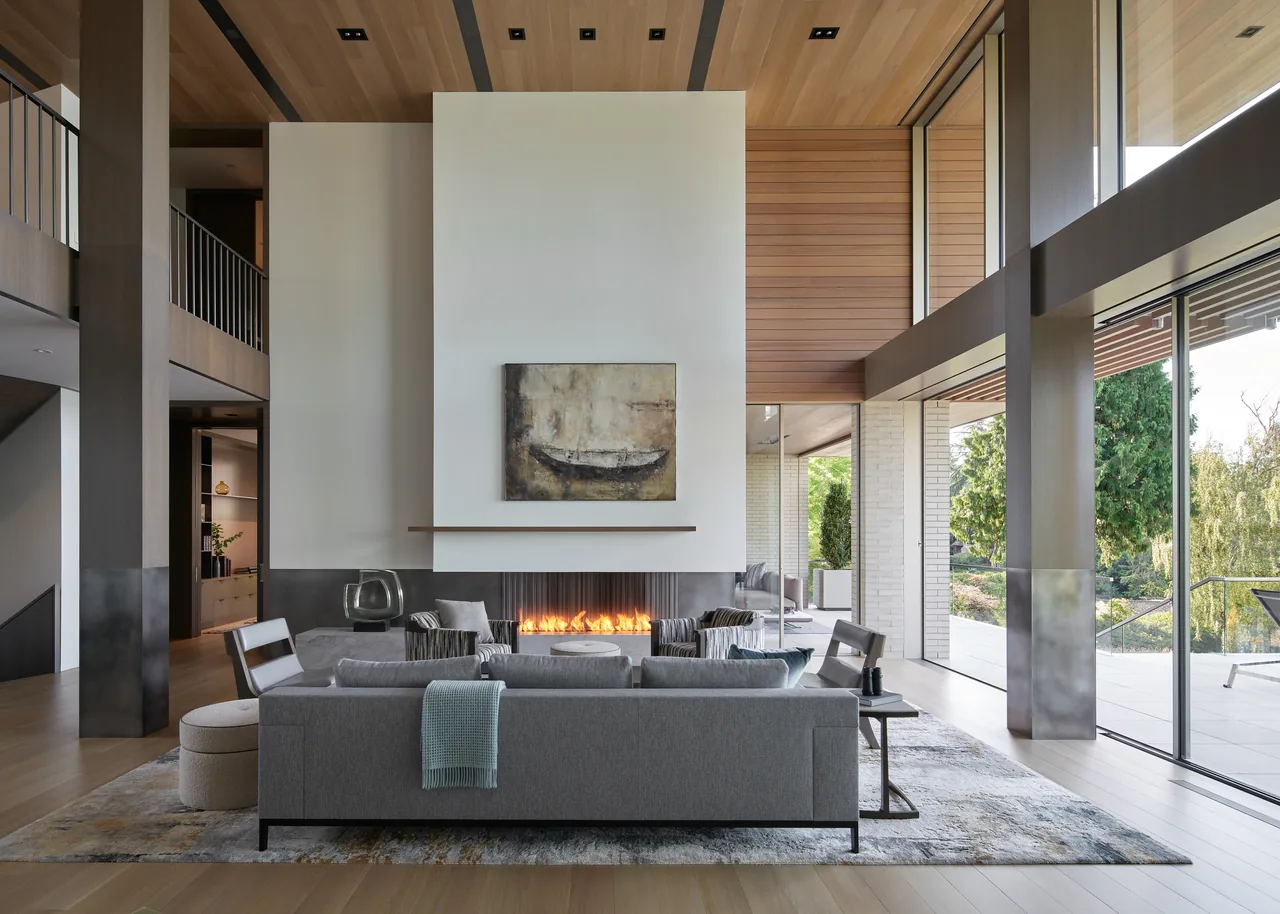 Spacious modern living room with a tall fireplace, floor-to-ceiling windows, and a high wood-paneled ceiling.