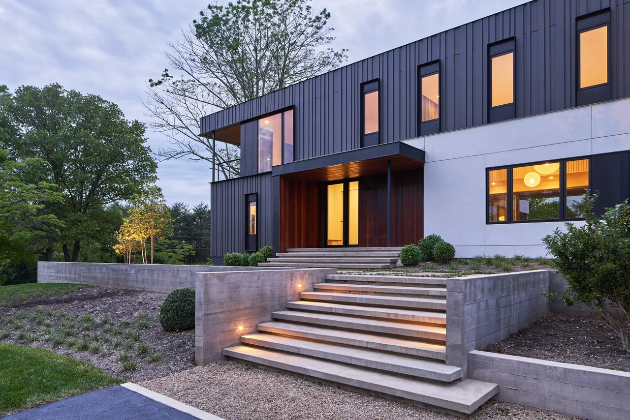 Modern two-story house exterior with black vertical metal siding, concrete stairs, and illuminated entry at dusk.