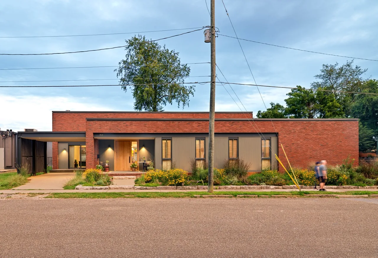 Single-story modern house with red brick and vertical wood siding exterior, flat roof, and front garden beds on a residential street.