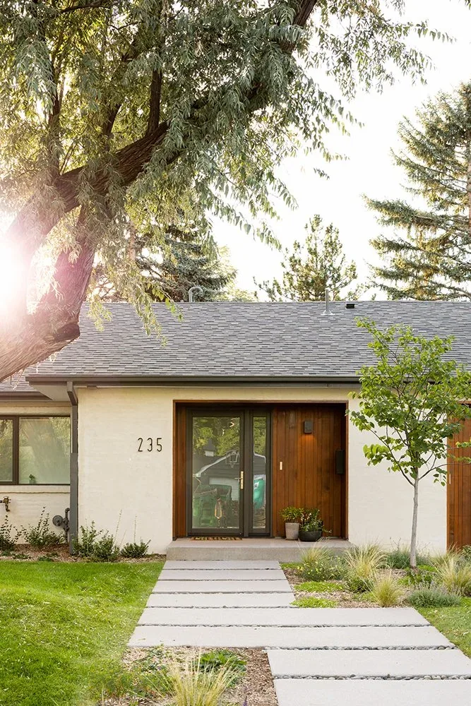 Front exterior of a modern house with a wood-paneled entrance and a concrete path leading to the door under sunlight through tree branches.