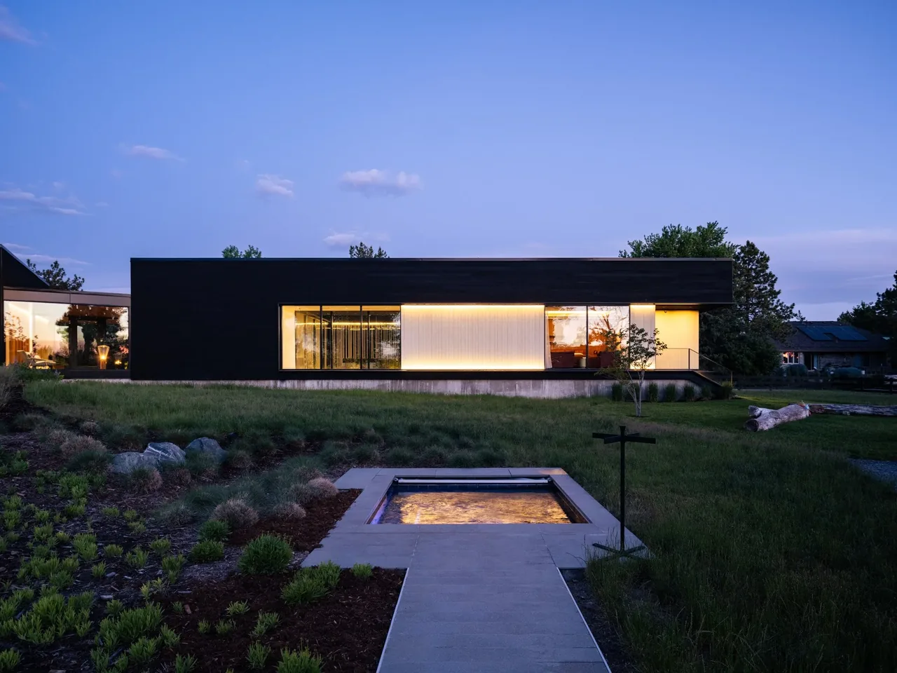 Modern rectangular house with dark exterior walls and large bright windows at dusk.