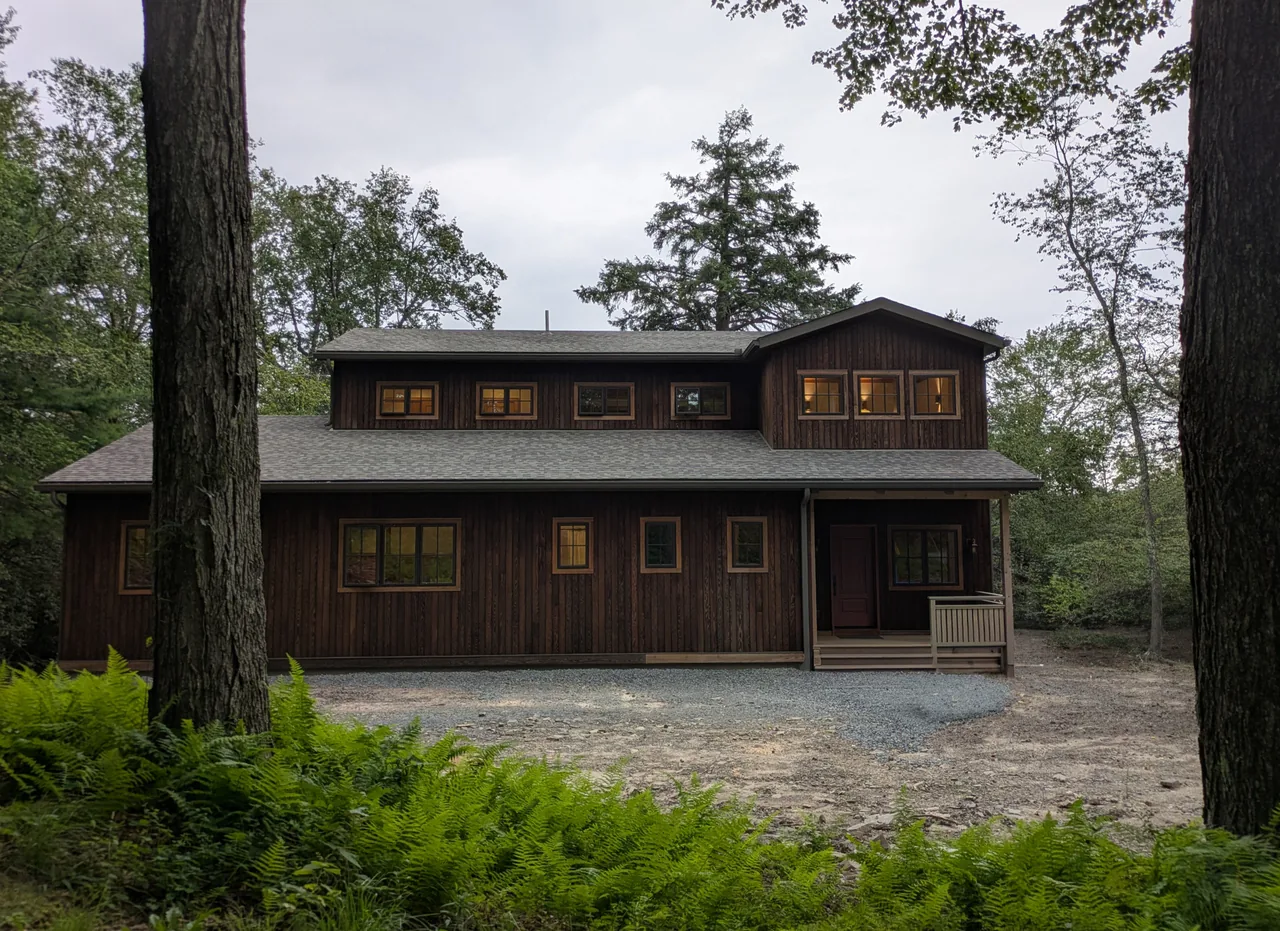 Two-story dark wood cottage in a forest clearing with several lit windows and a small covered porch.
