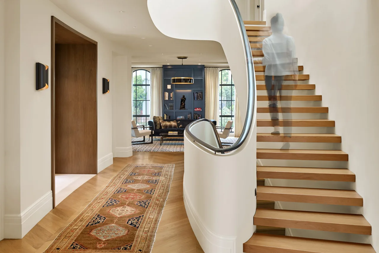 Curved wood floating staircase next to a hallway with a patterned runner leading to a living area with blue built-in shelving.