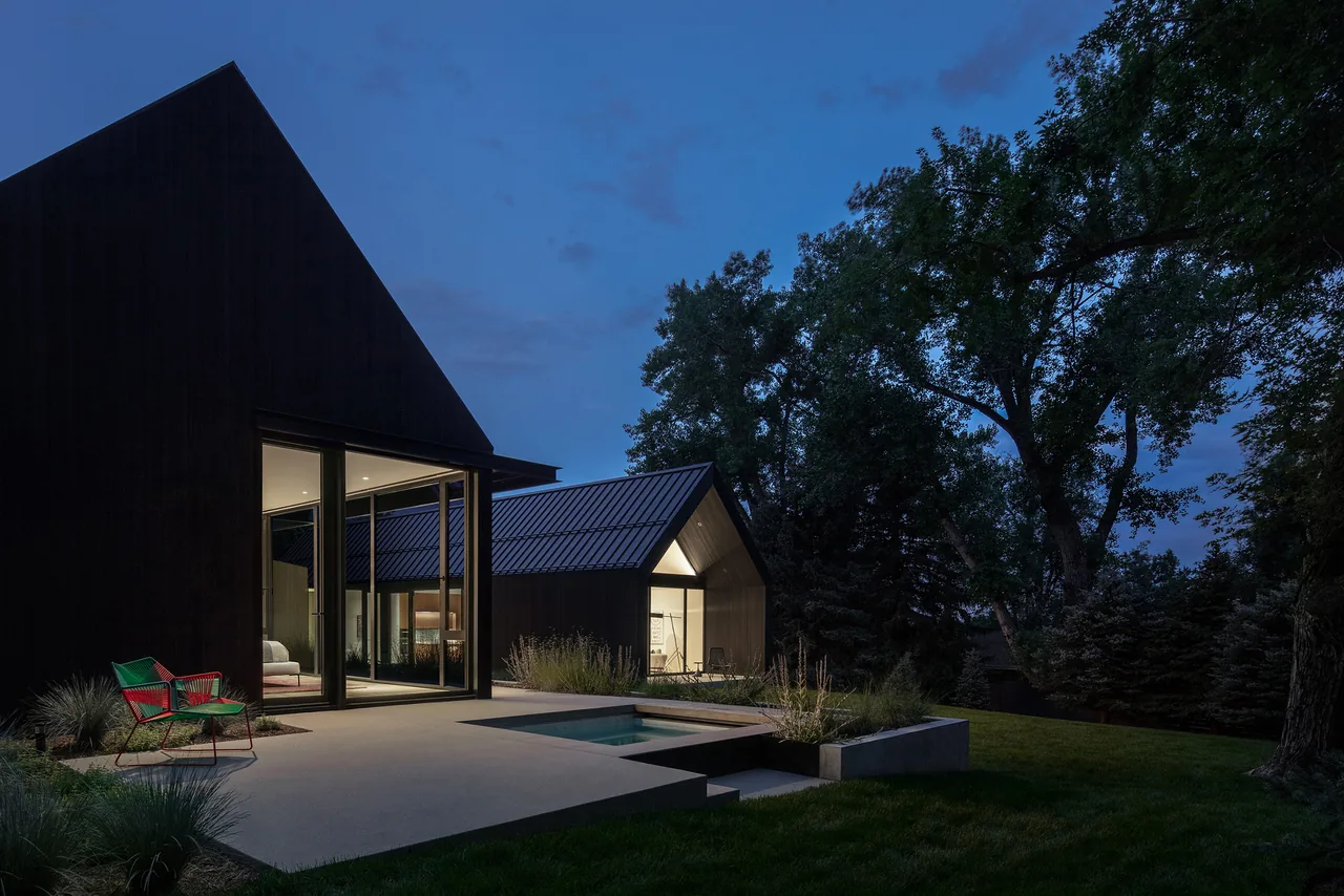 Night view of a modern dark house with three gabled roofs and a small pool in the backyard patio area surrounded by trees.