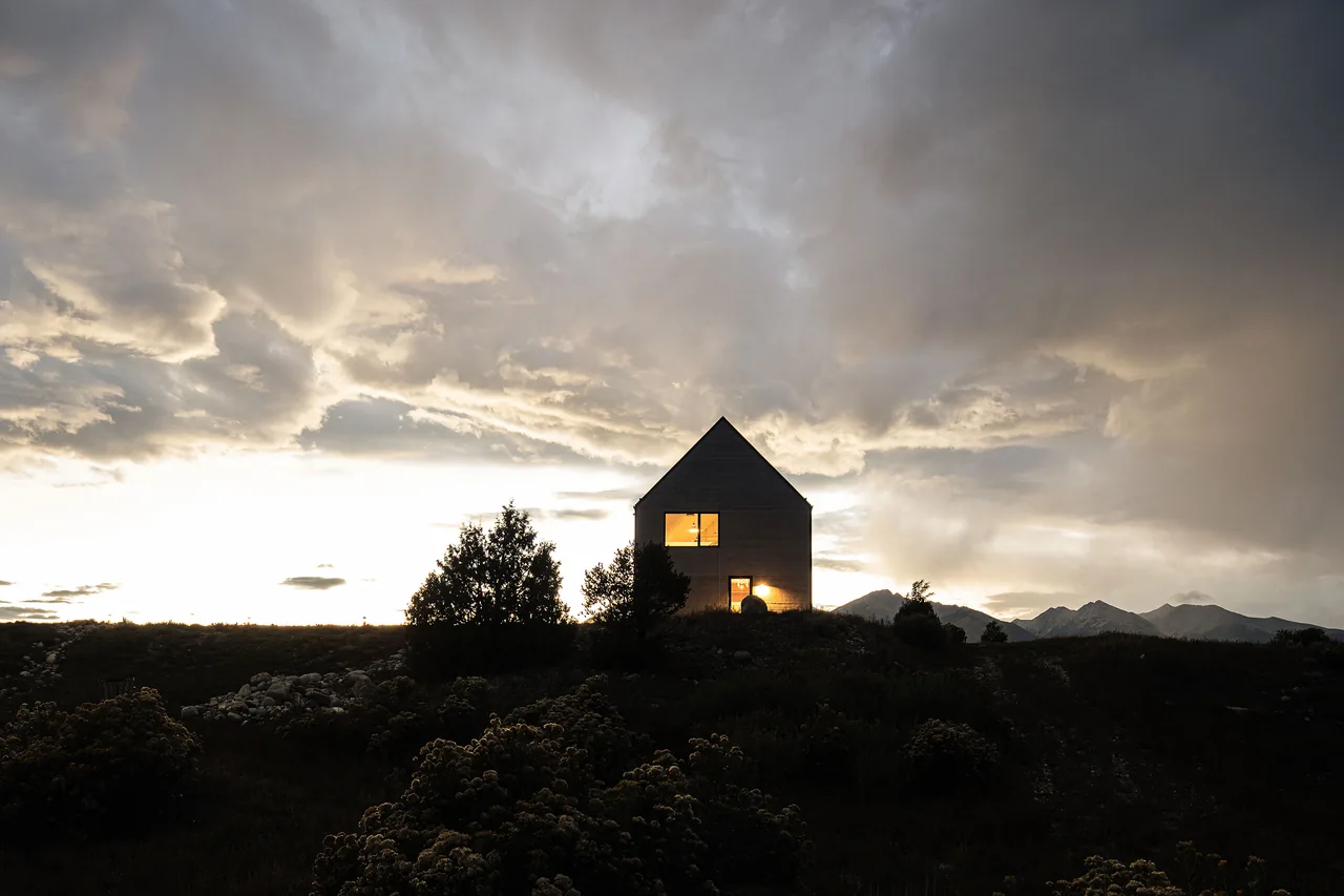Silhouetted house with pitched roof lit from inside at dusk on a hill surrounded by vegetation and mountains in the background.