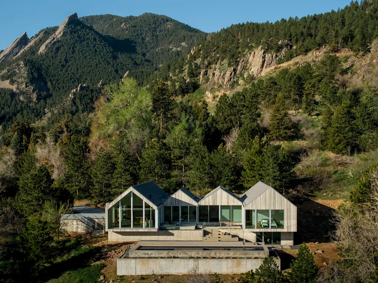 Modern house with four gabled sections and large windows nestled among pine trees and rocky hills.